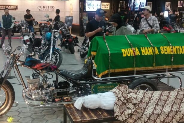 Custom motorcycle hearse on display with a green coffin cover reading 'Dunia Hanya Sementara' at an indoor bike show, with attendees in the background.