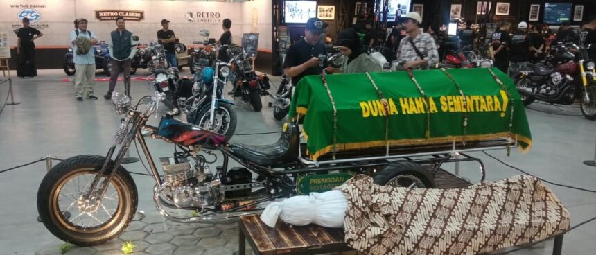 Custom motorcycle hearse on display with a green coffin cover reading 'Dunia Hanya Sementara' at an indoor bike show, with attendees in the background.