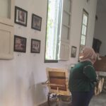 Woman wearing a hijab looks at framed photos on a white gallery wall with large windows and vintage furniture nearby.
