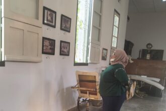 Woman wearing a hijab looks at framed photos on a white gallery wall with large windows and vintage furniture nearby.