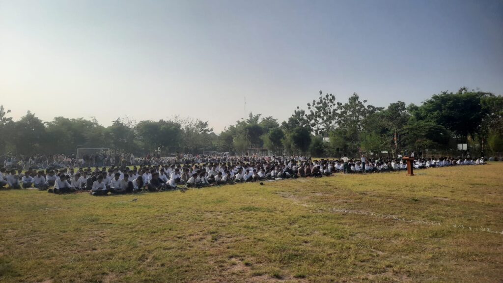 IMG 6708 - Mabur.co Large group of students in white shirts sitting on a grassy field for an outdoor assembly, with a podium to the right and trees in the background under a clear sky
