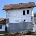 Old white two-story building with a red-tiled roof beside railway tracks, resting on a gray stone foundation.
