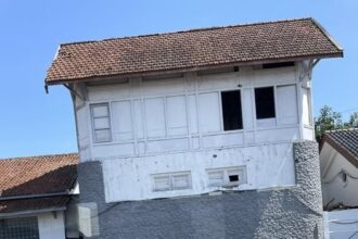 Old white two-story building with a red-tiled roof beside railway tracks, resting on a gray stone foundation.
