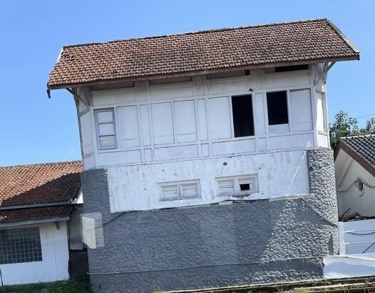 Old white two-story building with a red-tiled roof beside railway tracks, resting on a gray stone foundation.