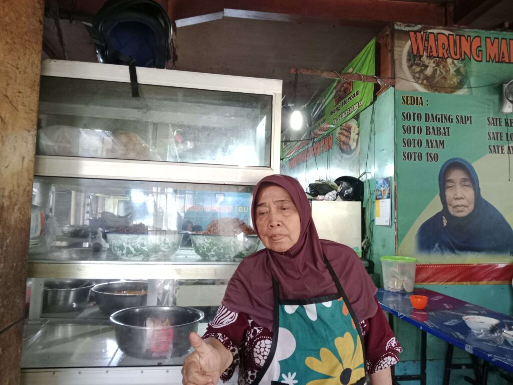 Woman in a maroon hijab and colorful apron stands behind a glass food counter at a street stall.