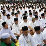 Large outdoor congregation of Muslim men in white shirts and black caps praying in unison on prayer mats.