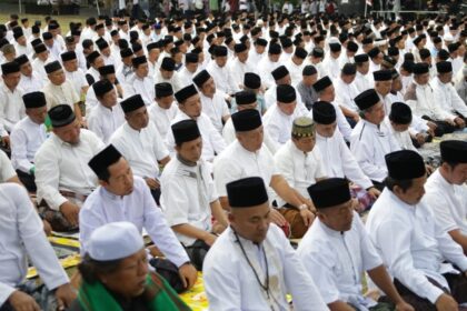 Large outdoor congregation of Muslim men in white shirts and black caps praying in unison on prayer mats.