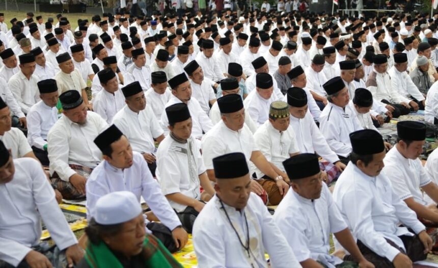 Large outdoor congregation of Muslim men in white shirts and black caps praying in unison on prayer mats.