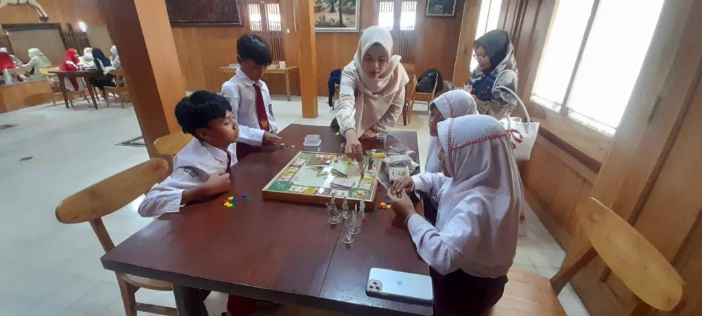 Group of children in uniforms playing a board game at a wooden table while adults supervise nearby in a bright room.