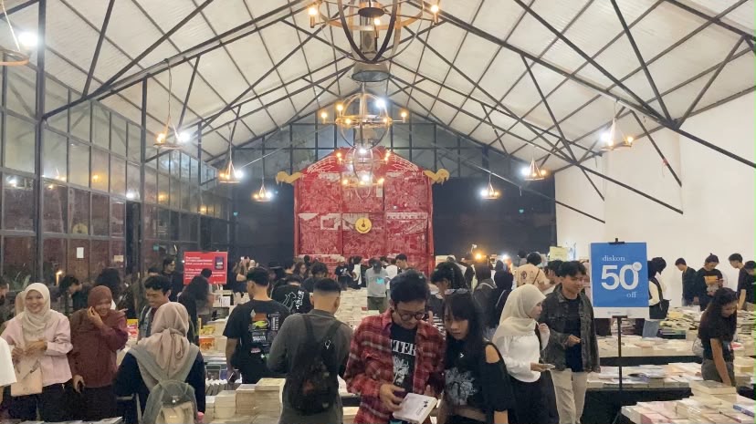 Crowd of people browsing books at a busy indoor book fair with a red stage backdrop.