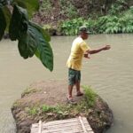 Man in a yellow shirt stands on a small rocky riverbank, arms extended as if fishing, with a bamboo raft docked nearby and green foliage overhead.