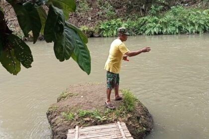 Man in a yellow shirt stands on a small rocky riverbank, arms extended as if fishing, with a bamboo raft docked nearby and green foliage overhead.