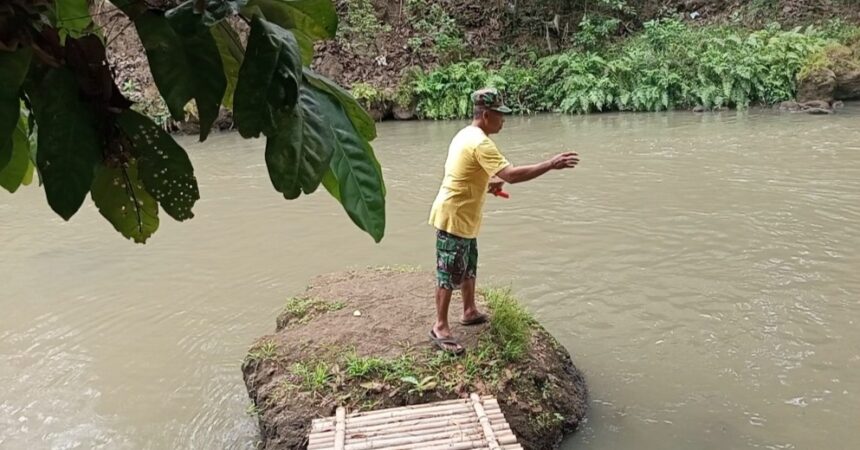 Man in a yellow shirt stands on a small rocky riverbank, arms extended as if fishing, with a bamboo raft docked nearby and green foliage overhead.