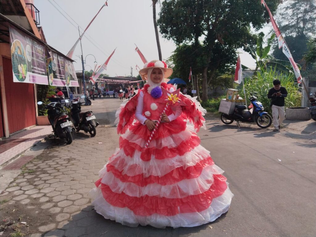 Person in a large pink and white ruffled dress with a wide straw hat posing in a street parade, holding a striped wand with a decorative flower.