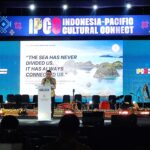 Speaker at a podium on stage with a large Indonesia-Pacific Cultural Connect banner above and audience silhouettes in front.