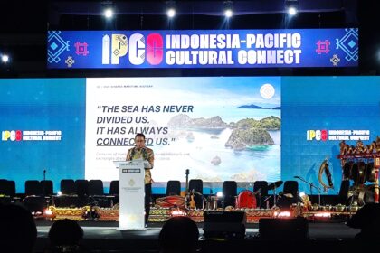 Speaker at a podium on stage with a large Indonesia-Pacific Cultural Connect banner above and audience silhouettes in front.