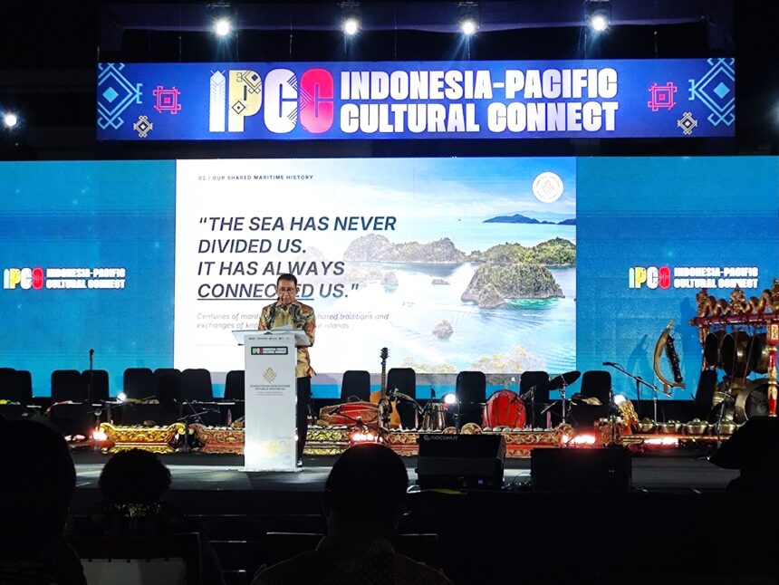 Speaker at a podium on stage with a large Indonesia-Pacific Cultural Connect banner above and audience silhouettes in front.