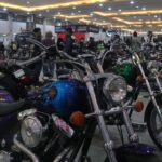 Indoor motorcycle show with a glossy blue cruiser in the foreground, chrome parts and round headlight, others lined up in the background.