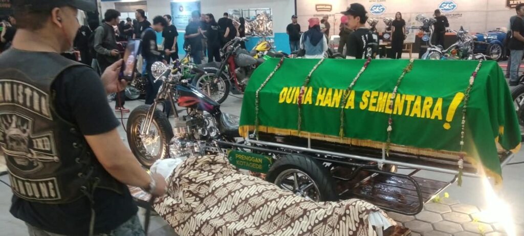 IMG 7890 - Mabur.co A body on a covered bier with a green banner reading'Dunia Hanya Sementara' at a motorcycle show as onlookers and bikes surround it.