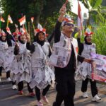 Parade participants in newspaper outfits march along a street, waving Indonesian flags and holding a banner reading Republik Indonesia.