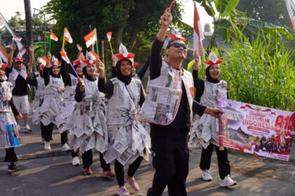 Parade participants in newspaper outfits march along a street, waving Indonesian flags and holding a banner reading Republik Indonesia.