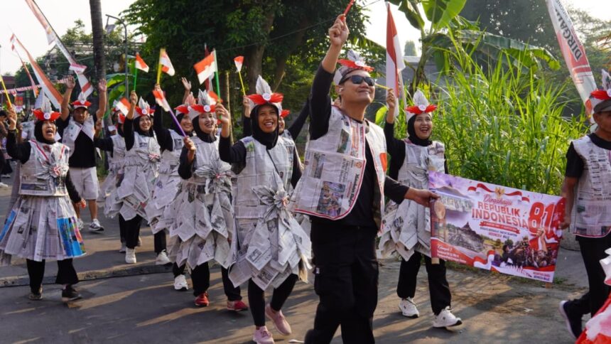 Parade participants in newspaper outfits march along a street, waving Indonesian flags and holding a banner reading Republik Indonesia.