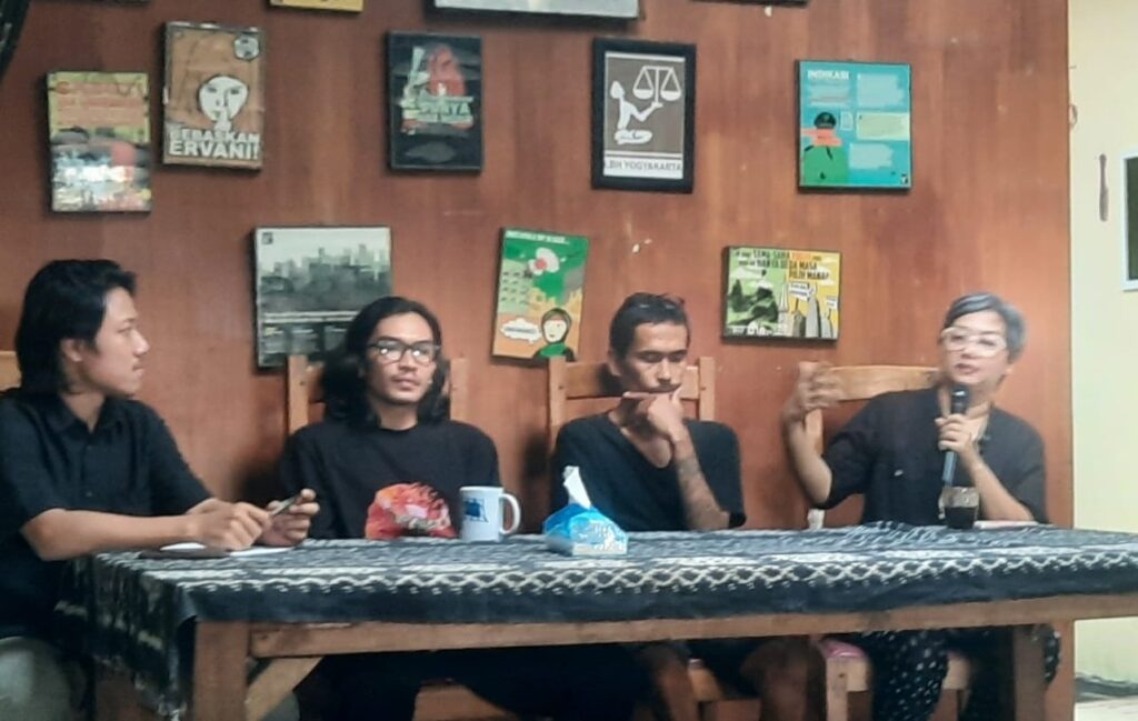 Panel discussion with four people seated at a wooden table; the person on the far right speaks into a microphone while others listen, wall with posters behind them.