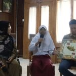 Three students sit indoors: a girl in a white hijab speaks into a microphone, flanked by a woman in a black hijab and a boy holding a boxed board game.