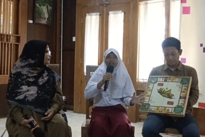 Three students sit indoors: a girl in a white hijab speaks into a microphone, flanked by a woman in a black hijab and a boy holding a boxed board game.