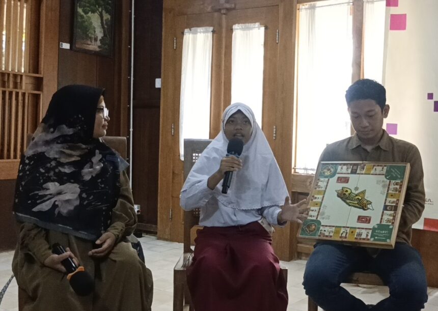 Three students sit indoors: a girl in a white hijab speaks into a microphone, flanked by a woman in a black hijab and a boy holding a boxed board game.