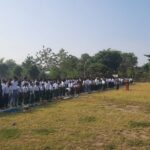 Large line of students in white shirts and dark pants standing on mats on a grassy school field during an outdoor assembly, with a speaker at a podium in the distance.
