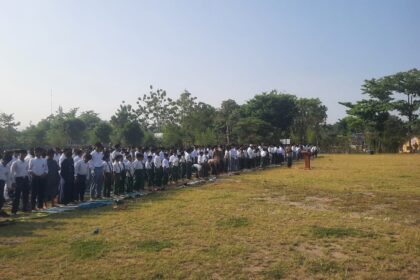 Large line of students in white shirts and dark pants standing on mats on a grassy school field during an outdoor assembly, with a speaker at a podium in the distance.