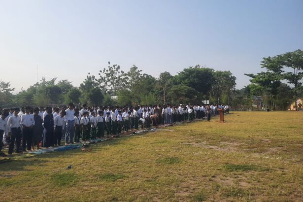 Large line of students in white shirts and dark pants standing on mats on a grassy school field during an outdoor assembly, with a speaker at a podium in the distance.