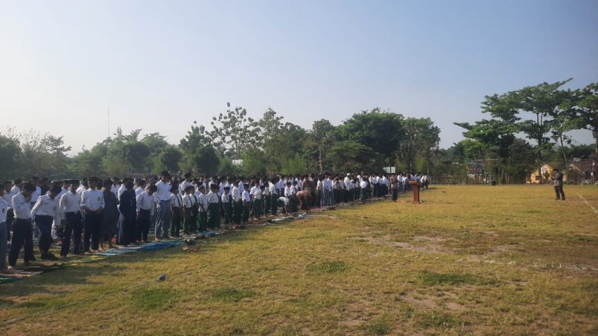 Large line of students in white shirts and dark pants standing on mats on a grassy school field during an outdoor assembly, with a speaker at a podium in the distance.