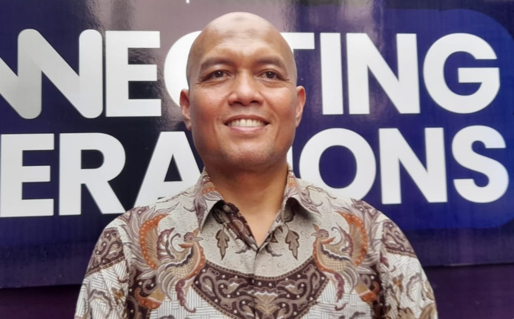 Smiling man in a patterned batik shirt stands in front of a dark blue banner with large white text.