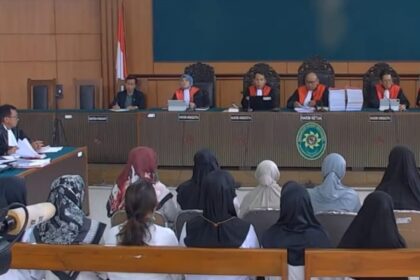 Courtroom scene with five judges in red robes seated at the bench, Indonesian flag behind them, audience watching.