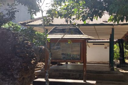 Wood-framed information board under a roofed structure in a shaded outdoor setting, with a stone wall to the left and trees overhead.
