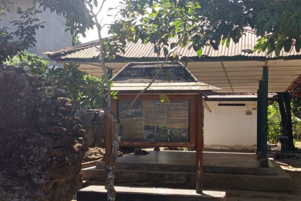 Wood-framed information board under a roofed structure in a shaded outdoor setting, with a stone wall to the left and trees overhead.