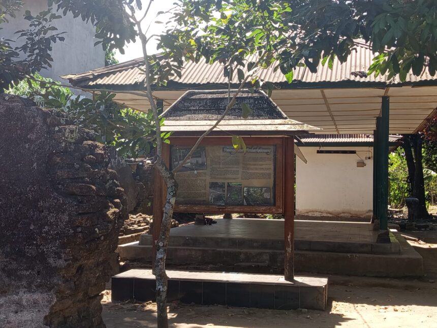 Wood-framed information board under a roofed structure in a shaded outdoor setting, with a stone wall to the left and trees overhead.