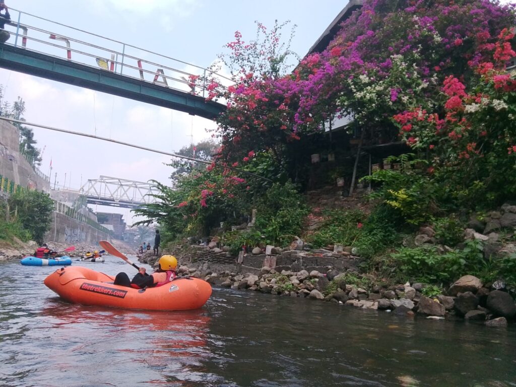 People wearing life jackets rafting on a calm river with an orange inflatable raft and rocky banks; colorful flowering vines cover the hillside on the right.