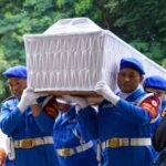 Uniformed pallbearers in blue carry a white coffin outdoors against a green, leafy background.