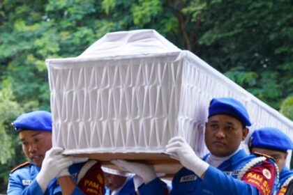 Uniformed pallbearers in blue carry a white coffin outdoors against a green, leafy background.