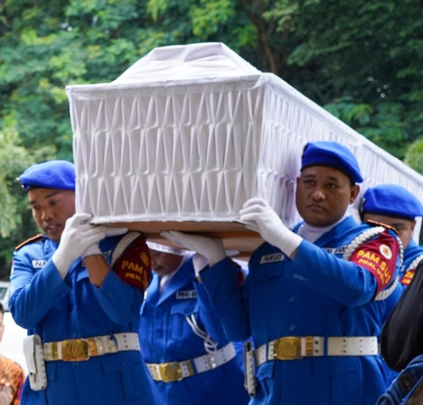 Uniformed pallbearers in blue carry a white coffin outdoors against a green, leafy background.
