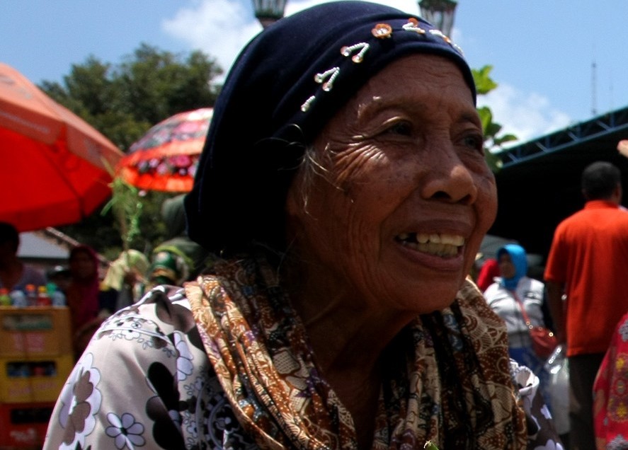 IMG 8907 - Mabur.co Smiling elderly woman wearing a dark headscarf and patterned blouse at an outdoor market with colorful umbrellas in the background.