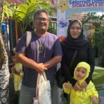 Man and woman with a young girl in yellow uniforms posing in front of a colorful school welcome banner reading 'Selamat Datang Siswa MPLS 2026'