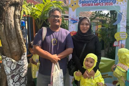 Man and woman with a young girl in yellow uniforms posing in front of a colorful school welcome banner reading 'Selamat Datang Siswa MPLS 2026'