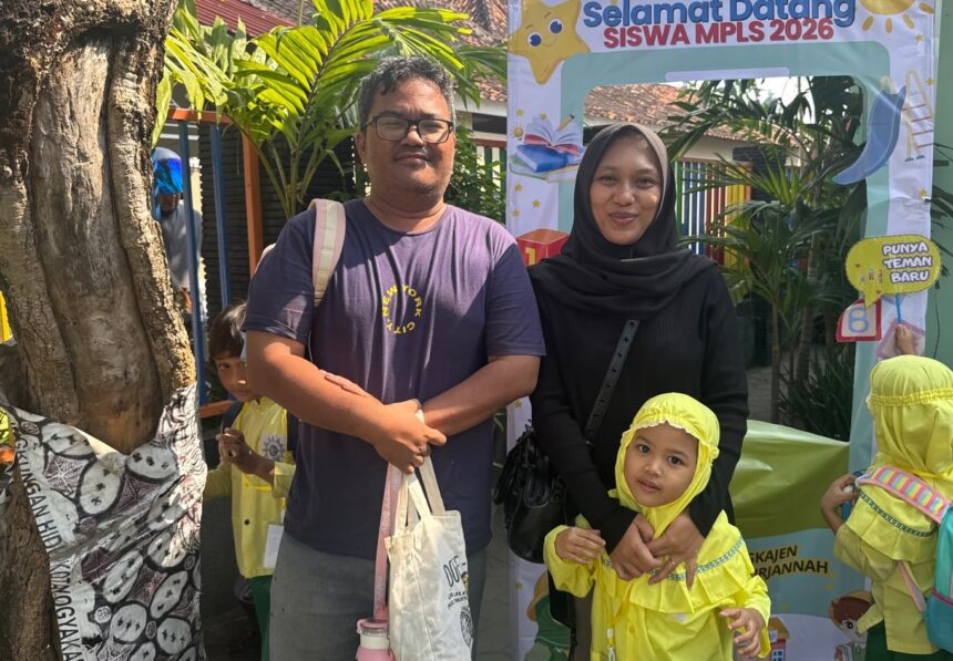 Man and woman with a young girl in yellow uniforms posing in front of a colorful school welcome banner reading 'Selamat Datang Siswa MPLS 2026'