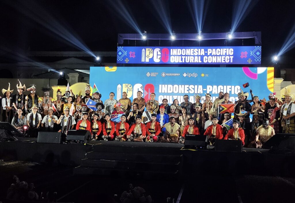 Group of performers in colorful traditional costumes posing on a stage at night, with Indonesia-Pacific Cultural Connect banners behind.