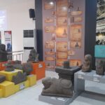 Display of various stone statues and carvings arranged on orange and yellow pedestals in a museum gallery, with informational plaques nearby.