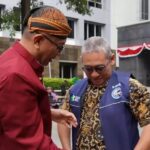 Two men in an outdoor plaza share a moment, one in a maroon shirt and patterned headwrap, the other in a blue vest and batik shirt, looking at the other's hand.